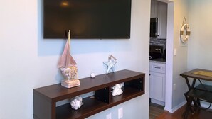 A living room with a flat-screen TV mounted on the wall, a wooden console table with decorative items, and a kitchen area visible through an open doorway. - Pearl of The Sea, 1 BDRM Condo, 2nd floor, heated pool, 2 min walk to the beach  (Madeira Beach)