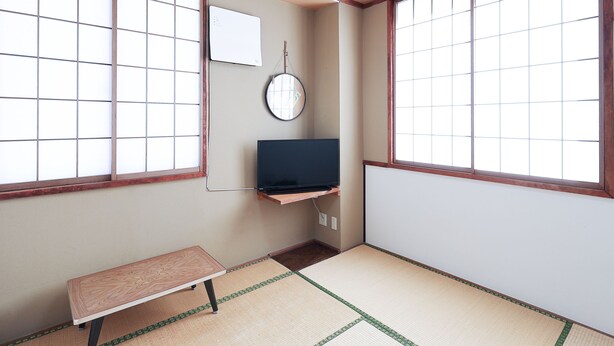 Una habitación japonesa tradicional con suelo de tatami, una mesa baja, un televisor sobre una estantería y ventanas amplias con marcos de madera.
