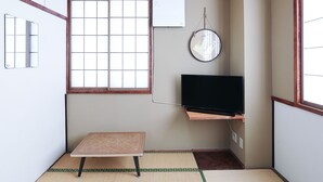 Una habitación pequeña con suelo de tatami, una mesa de madera baja, un televisor sobre un soporte de madera y una ventana con diseño de cuadrícula.
