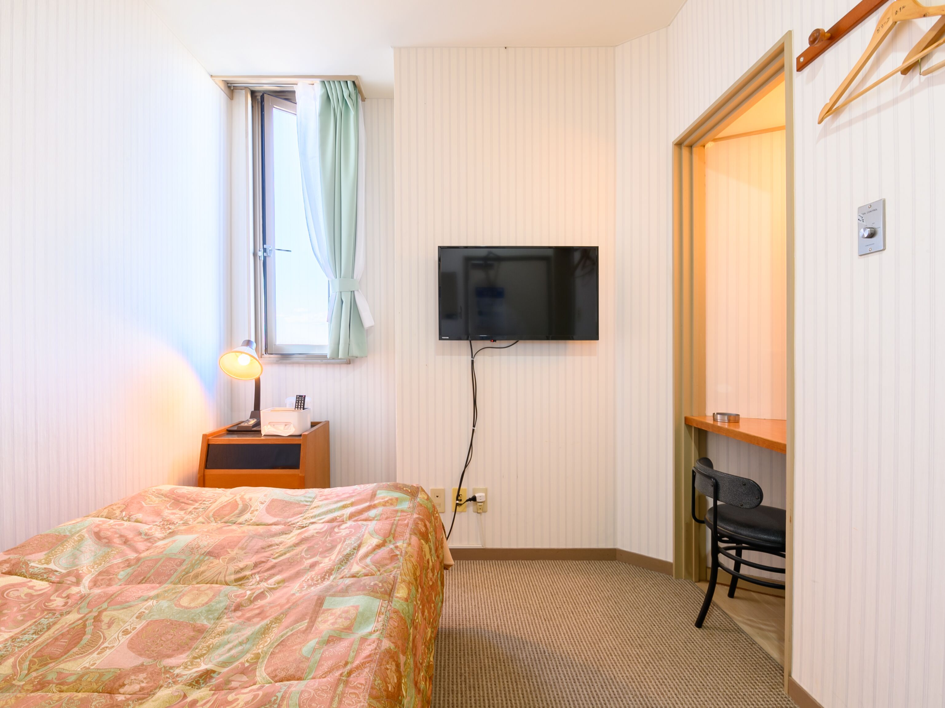 A hotel room with a bed, a desk with a lamp, a television mounted on the wall, and a chair.
