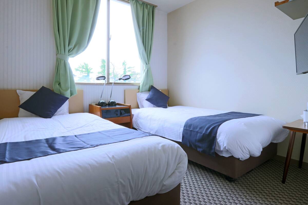 Habitación de hotel con dos camas individuales, una mesita de noche con lámpara, una ventana con cortinas y una pequeña mesa con un florero.