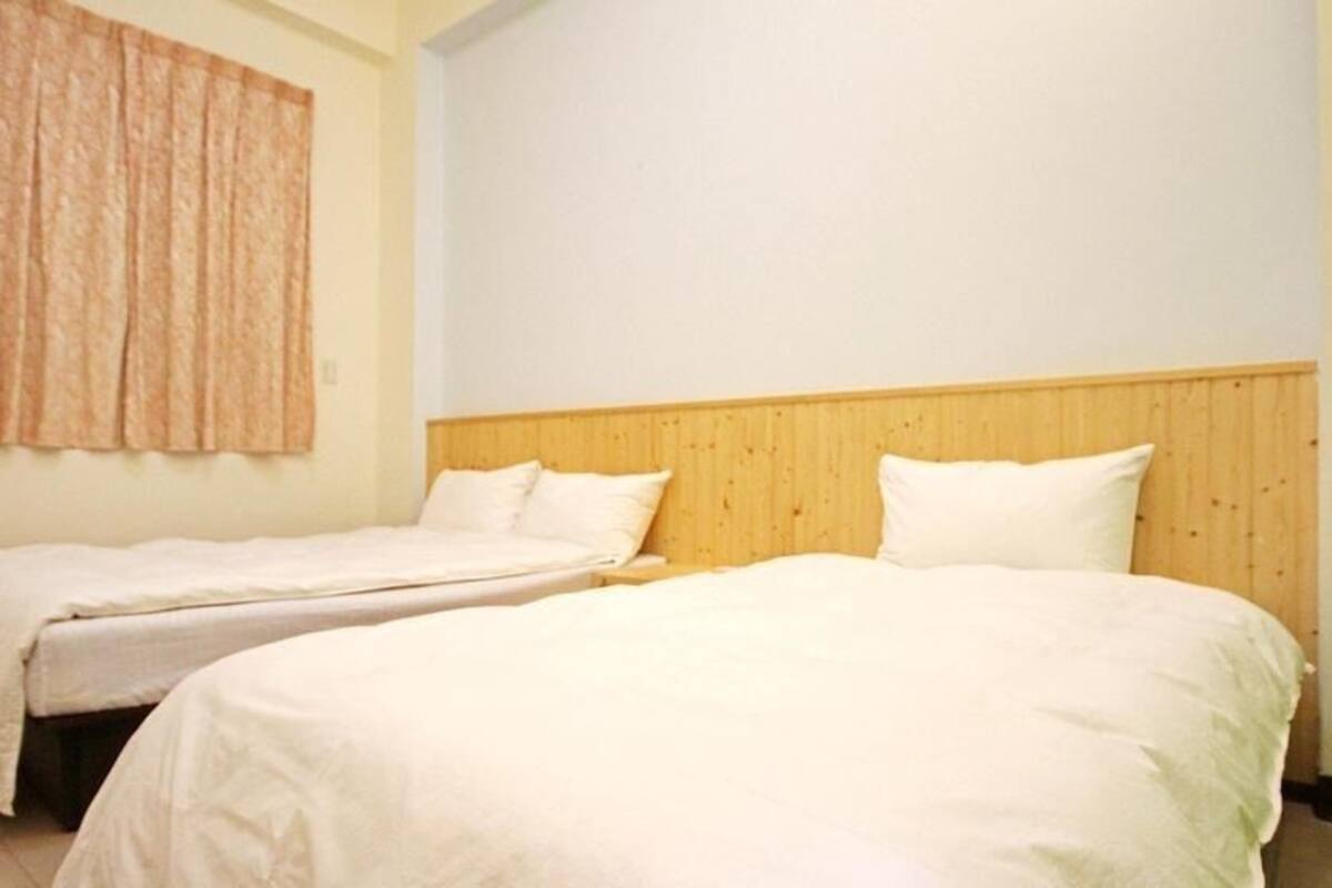 Une chambre avec deux lits simples, une tête de lit en bois et une grande fenêtre avec des rideaux.