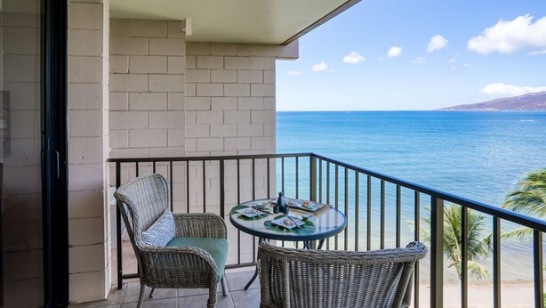 A balcony with wicker chairs and a table, overlooking a beach and ocean.