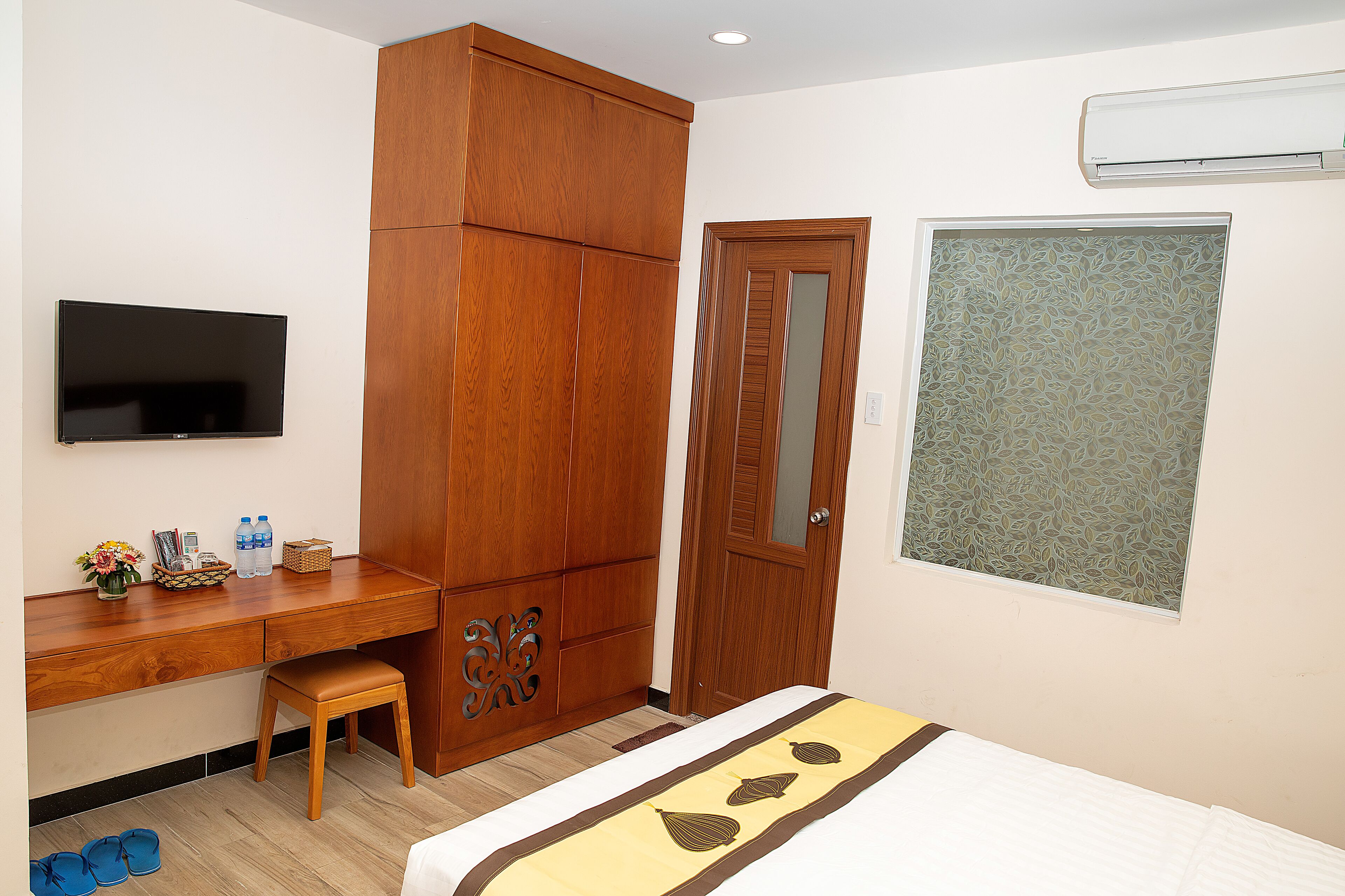 A hotel room with a wooden wardrobe, a desk with a flat-screen TV, a bed with a decorative bedspread, and a window with a leaf-patterned blind.