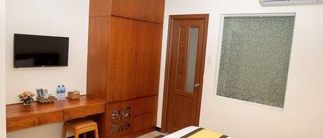 A hotel room with a wooden wardrobe, a desk with a flat-screen TV, a bed with a decorative bedspread, and a window with a leaf-patterned blind.