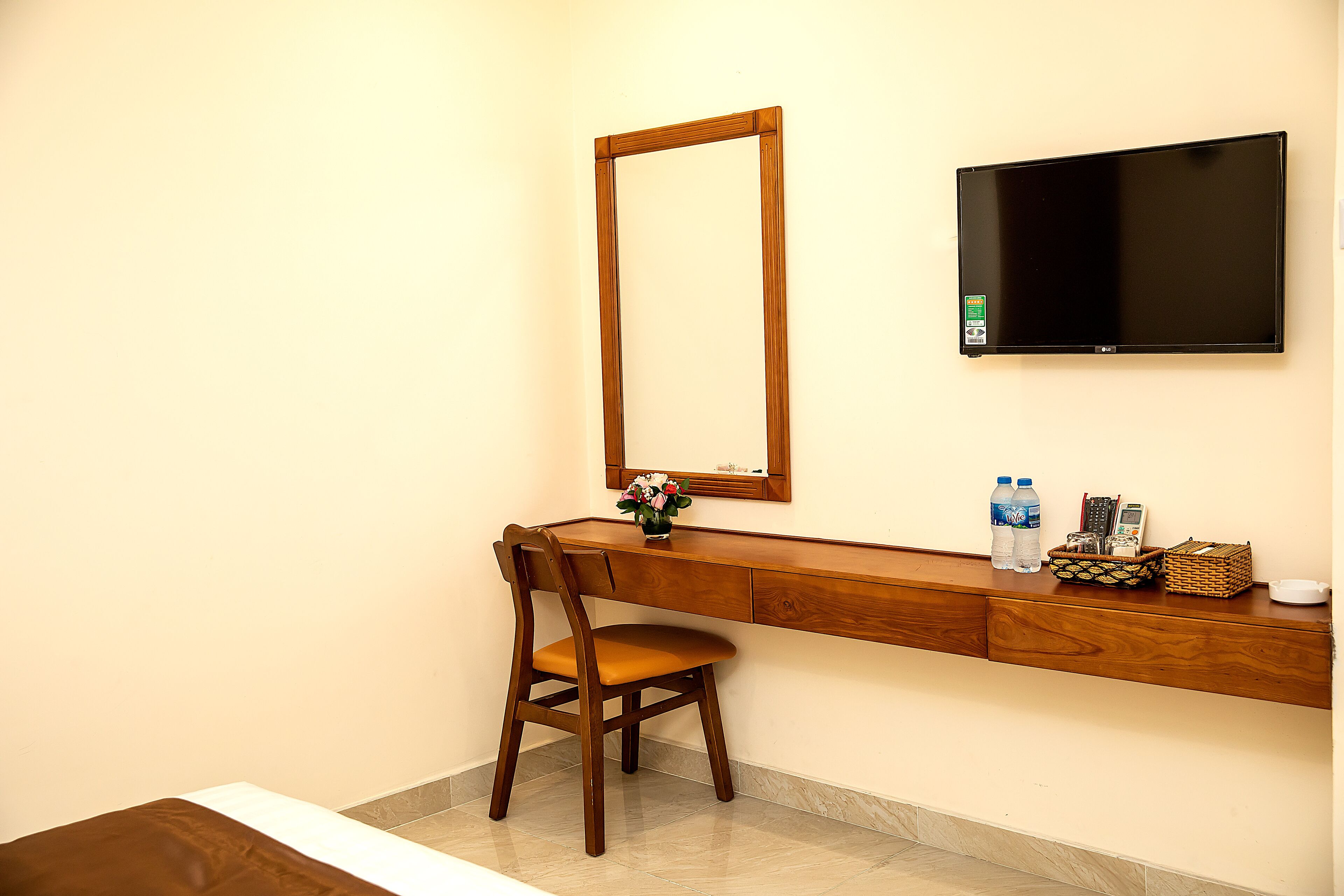 A hotel room with a wooden desk, a chair, a flat-screen TV, a mirror, and a small shelf with toiletries.
