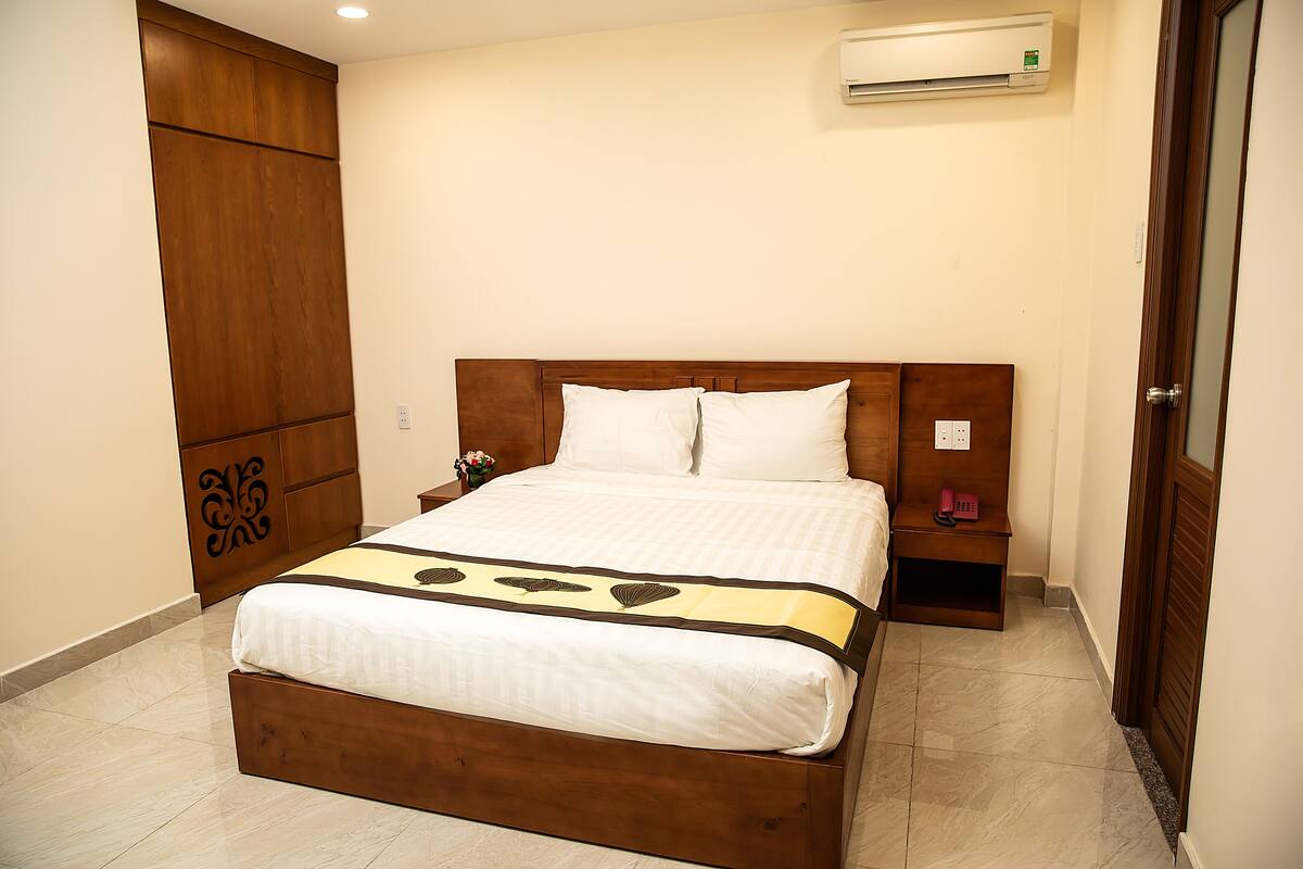 Una cama bien hecha con cabecera de madera, una mesita de noche con un teléfono y un equipo de aire acondicionado en la pared.