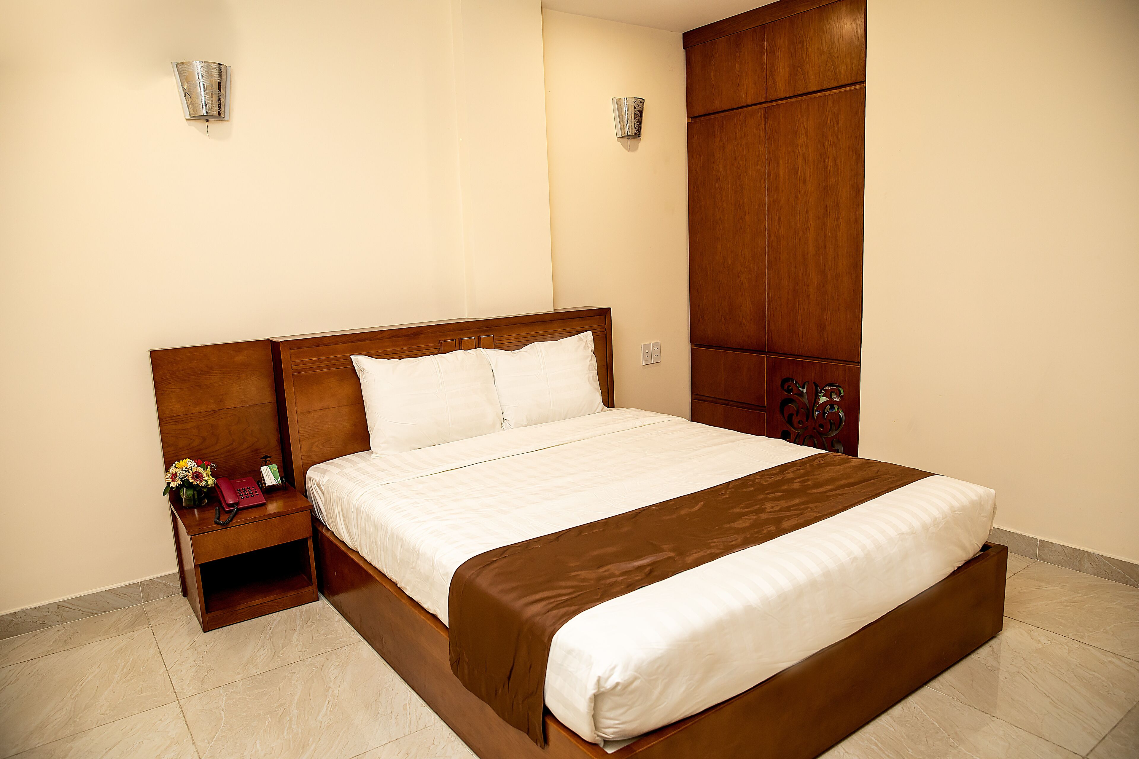 A hotel room with a large bed, wooden headboard, bedside table with a phone and flowers, and a wardrobe.