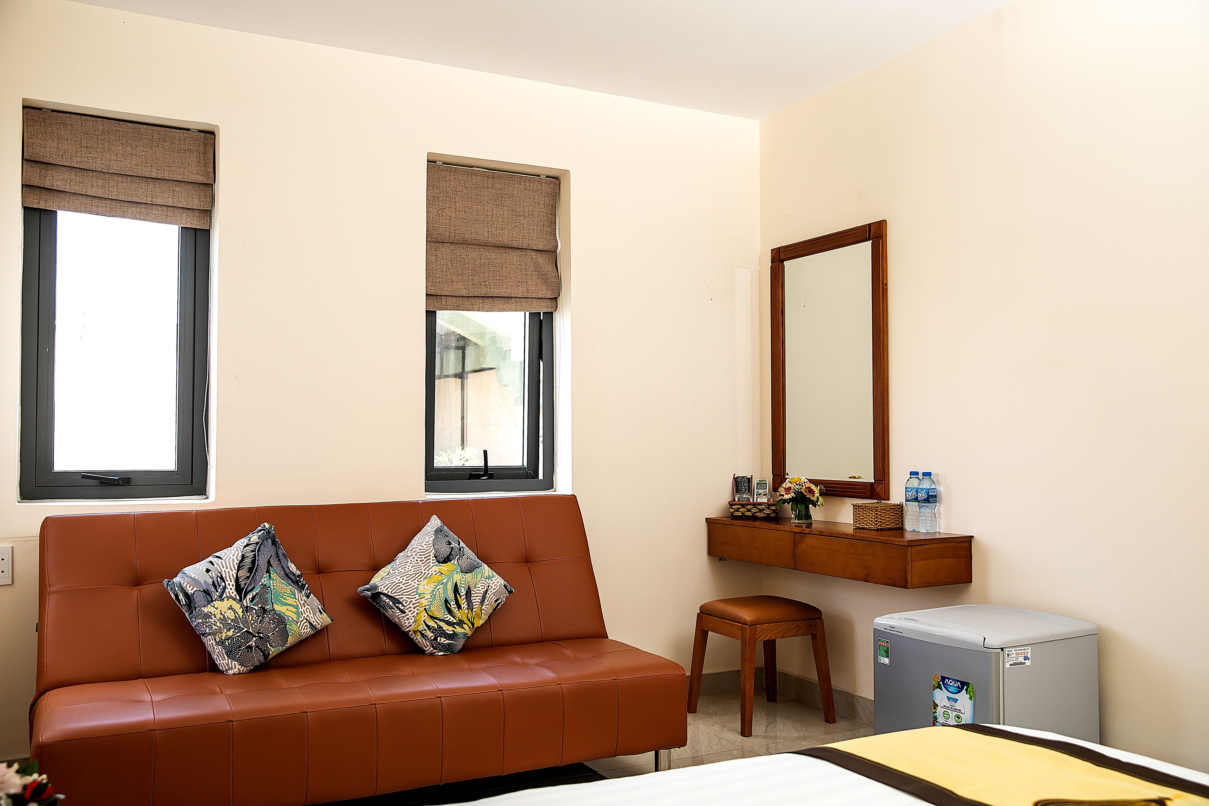 Habitación de hotel con un sofá de cuero marrón, dos almohadones decorativos, un taburete de madera, un refrigerador compacto, un espejo y una ventana con persiana.