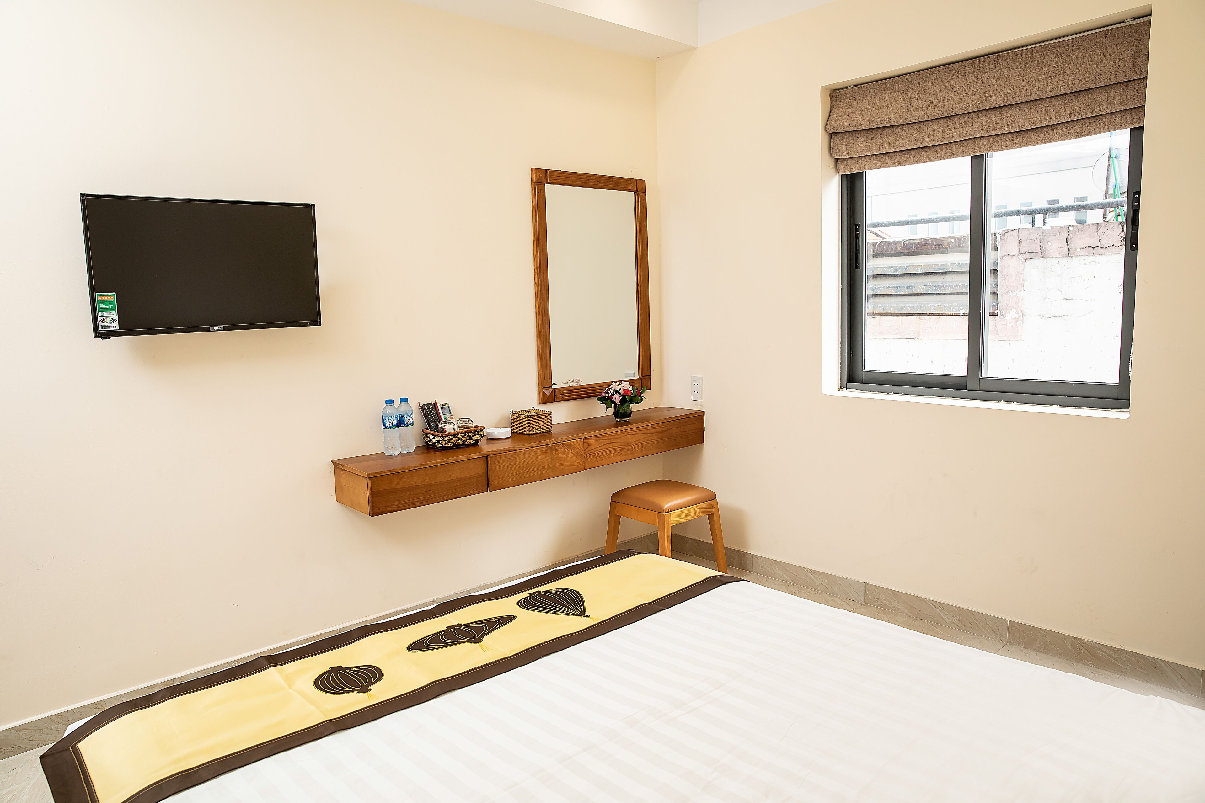 A hotel room with a bed, a TV mounted on the wall, a wooden bench, a mirror, and a window with a view of the outside.