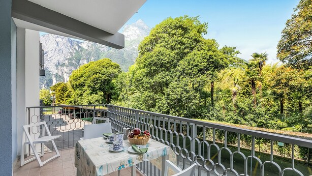Un balcón con mesa lista para comer, rodeado de barandilla, con vista a un jardín exuberante y una montaña.