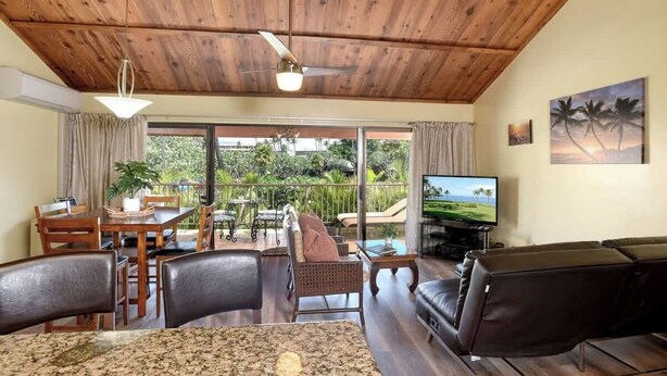 A spacious living area with a wooden ceiling, a television, a dining table with chairs, and a view of the outdoors. - Spacious Koa Resort Condo, Across Street From Beac (Kihei)