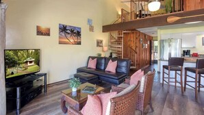 A spacious living room with a black leather sofa, a wooden coffee table, and a TV on a stand. A staircase leads to an upper level with a wooden railing. - Spacious Koa Resort Condo, Across Street From Beac (Kihei)