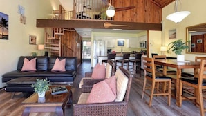 A spacious living area with a black leather sofa, wicker chairs, and a wooden dining table. A staircase leads to an upper level with a balcony. - Spacious Koa Resort Condo, Across Street From Beac (Kihei)