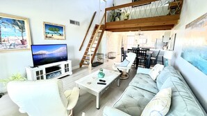Amplia sala de estar con televisión, asientos cómodos, una escalera y una cocina al fondo.