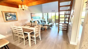 Amplia sala de estar con mesa de comedor, sillas, sofá y mesa de centro. Se aprecia una araña, un cuadro y vistas al exterior.