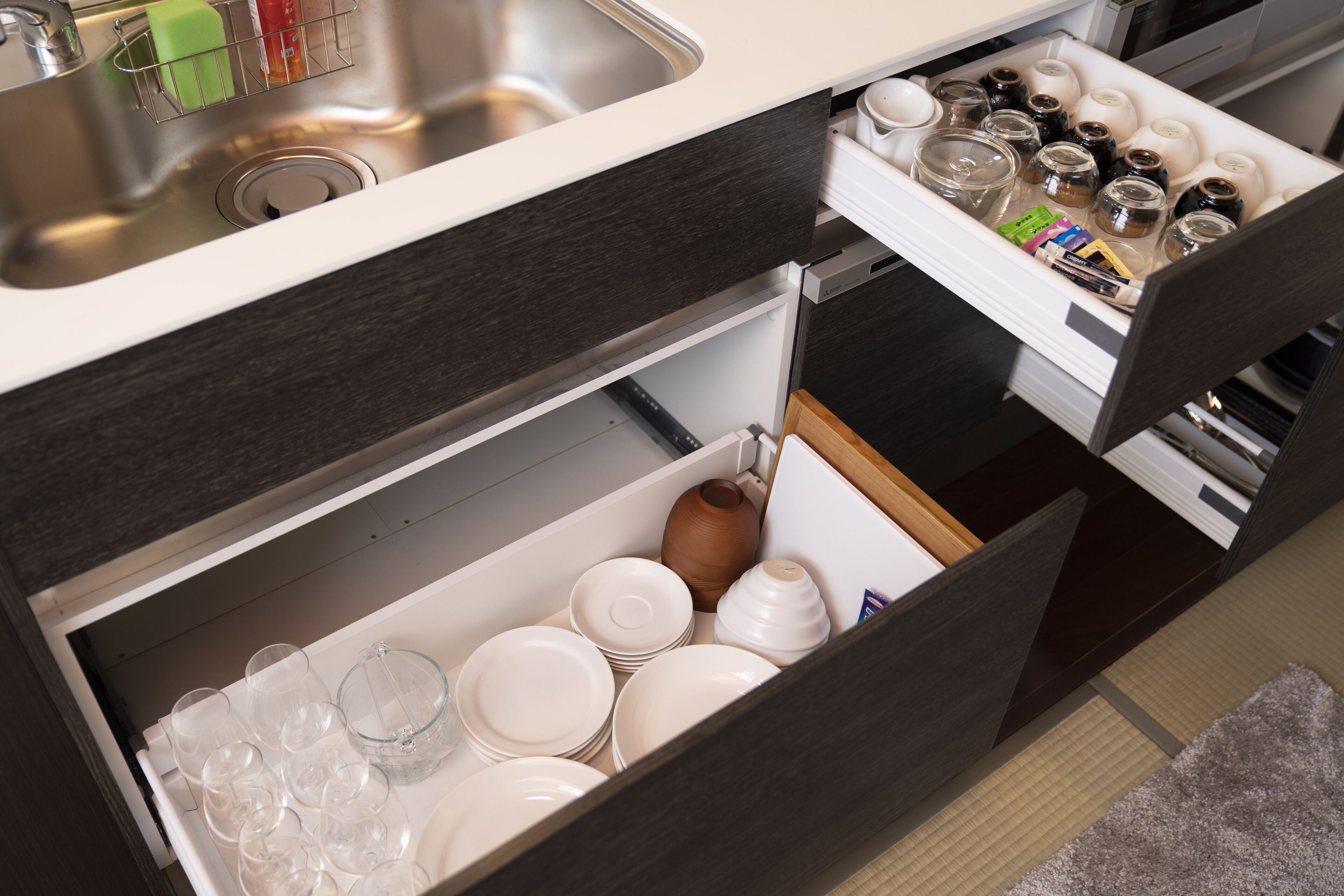 A kitchen with a sink, a countertop, and a drawer containing plates, glasses, and jars.
