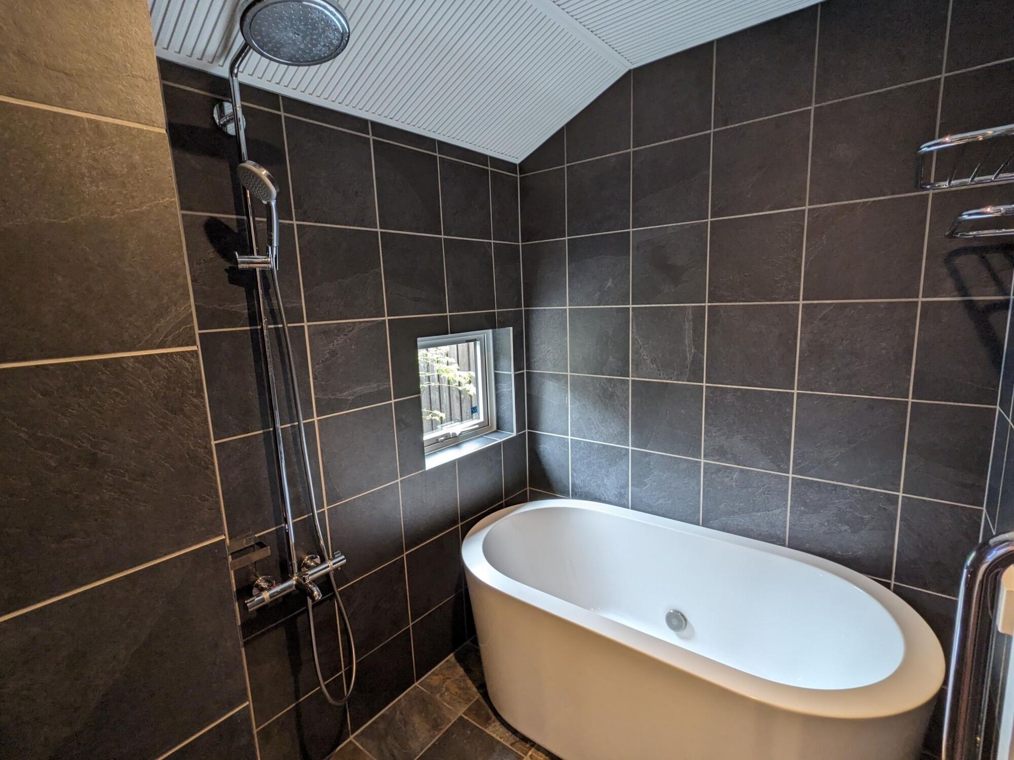Un baño moderno con baldosas oscuras, una bañera independiente, un cabezal de ducha y una ventana.