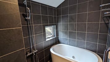 Un baño moderno con baldosas oscuras, una bañera independiente, un cabezal de ducha y una ventana.