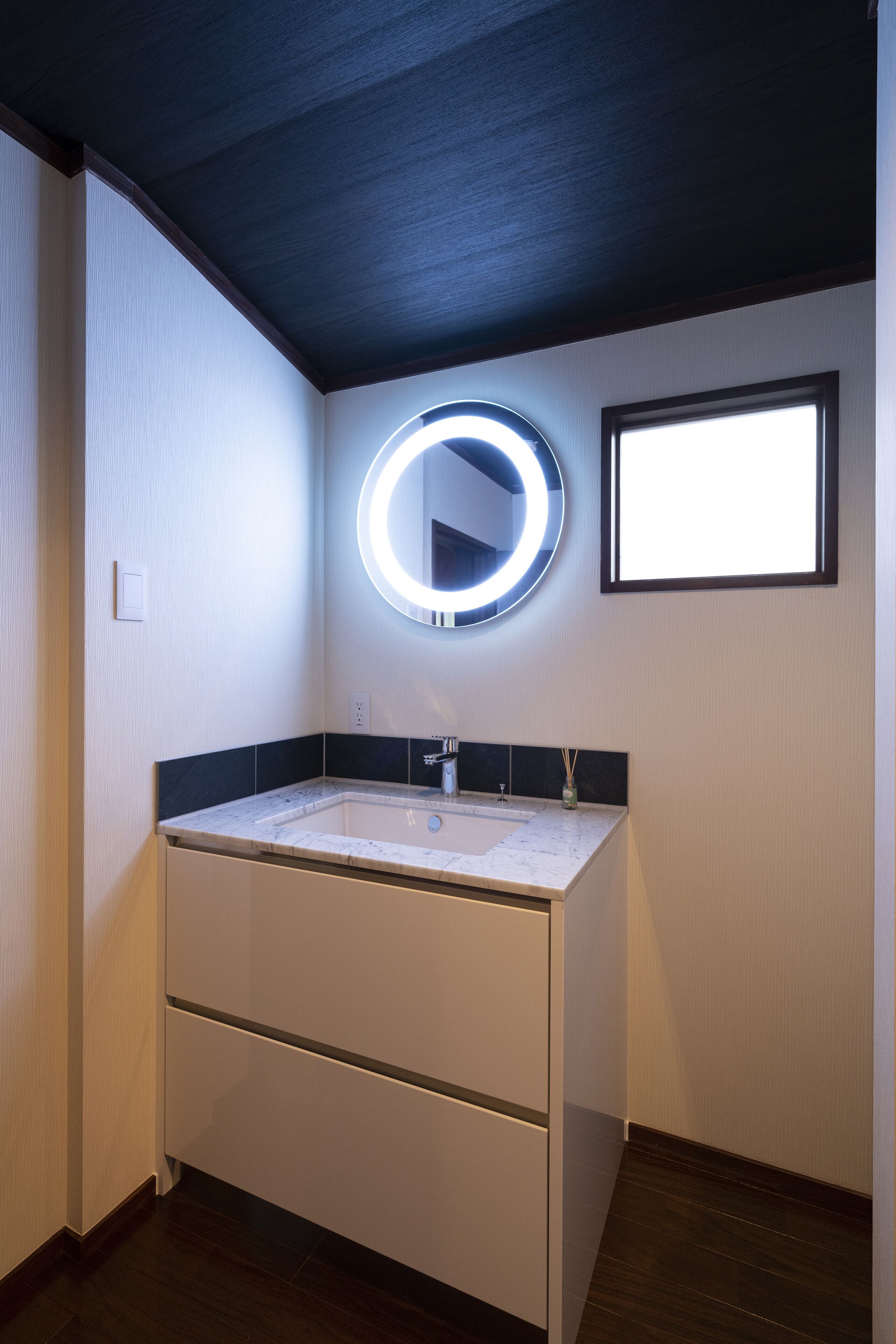 A bathroom with a white vanity, a round mirror with a light, and a window.