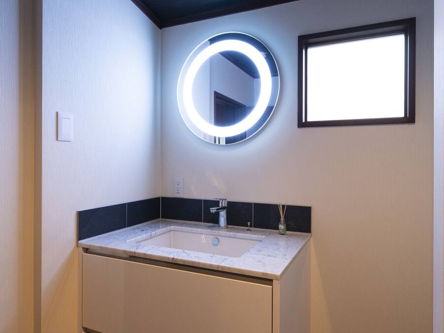 A bathroom with a white vanity, a round mirror with a light, and a window.