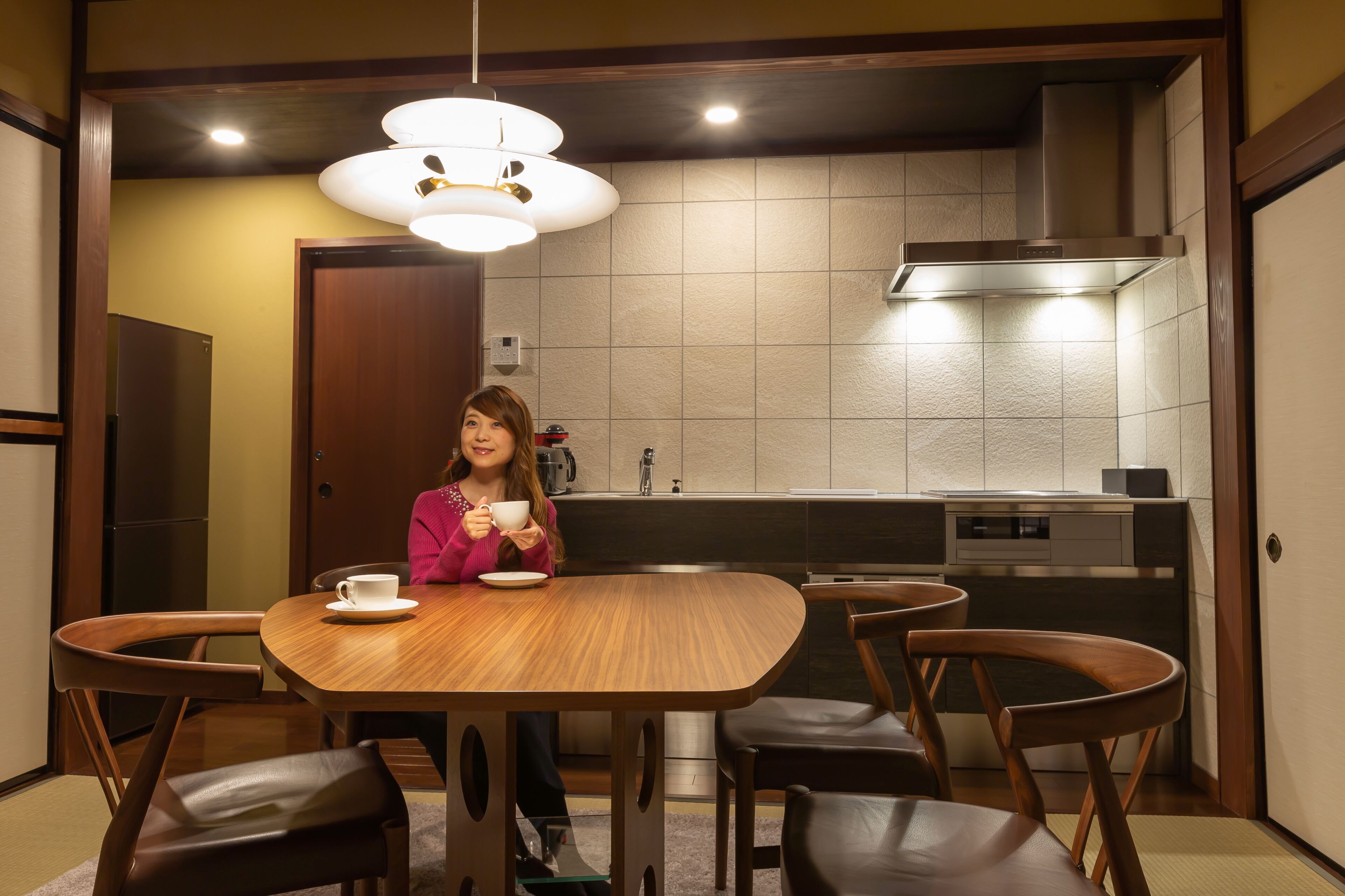 Un coin repas avec une table et des chaises en bois, une personne tenant une tasse, et une cuisine en arrière-plan.