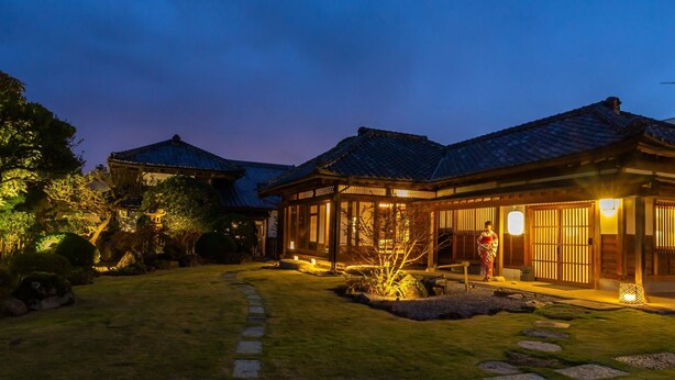 A traditional Japanese-style building with a tiled roof and wooden structure, surrounded by a well-maintained garden with stone pathways and a small tree. - Iizukatei (Nakagawa)
