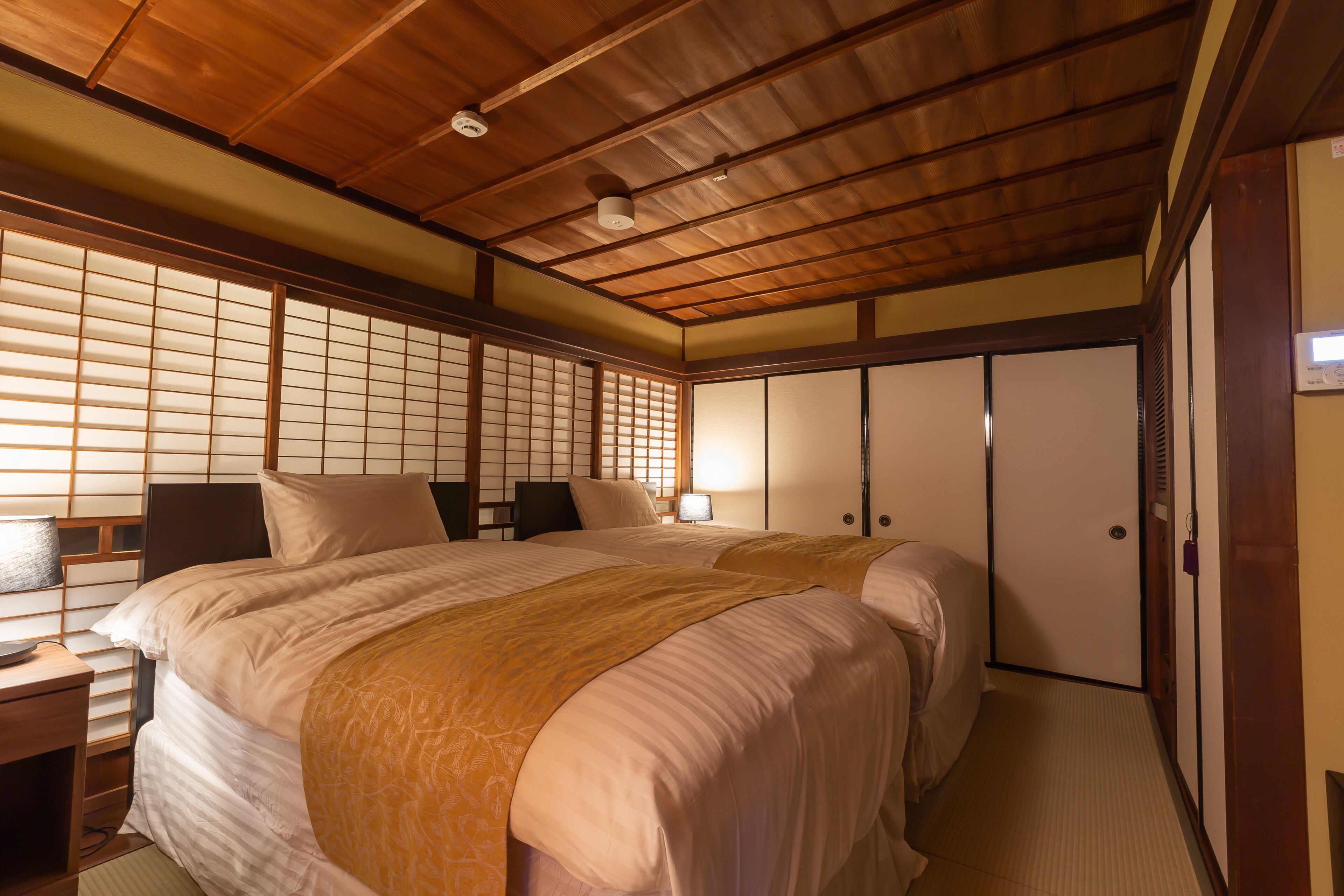 A traditional Japanese-style room with tatami flooring, a low wooden ceiling, and sliding shoji doors. There are two beds with beige bedding and a small table with a lamp.
