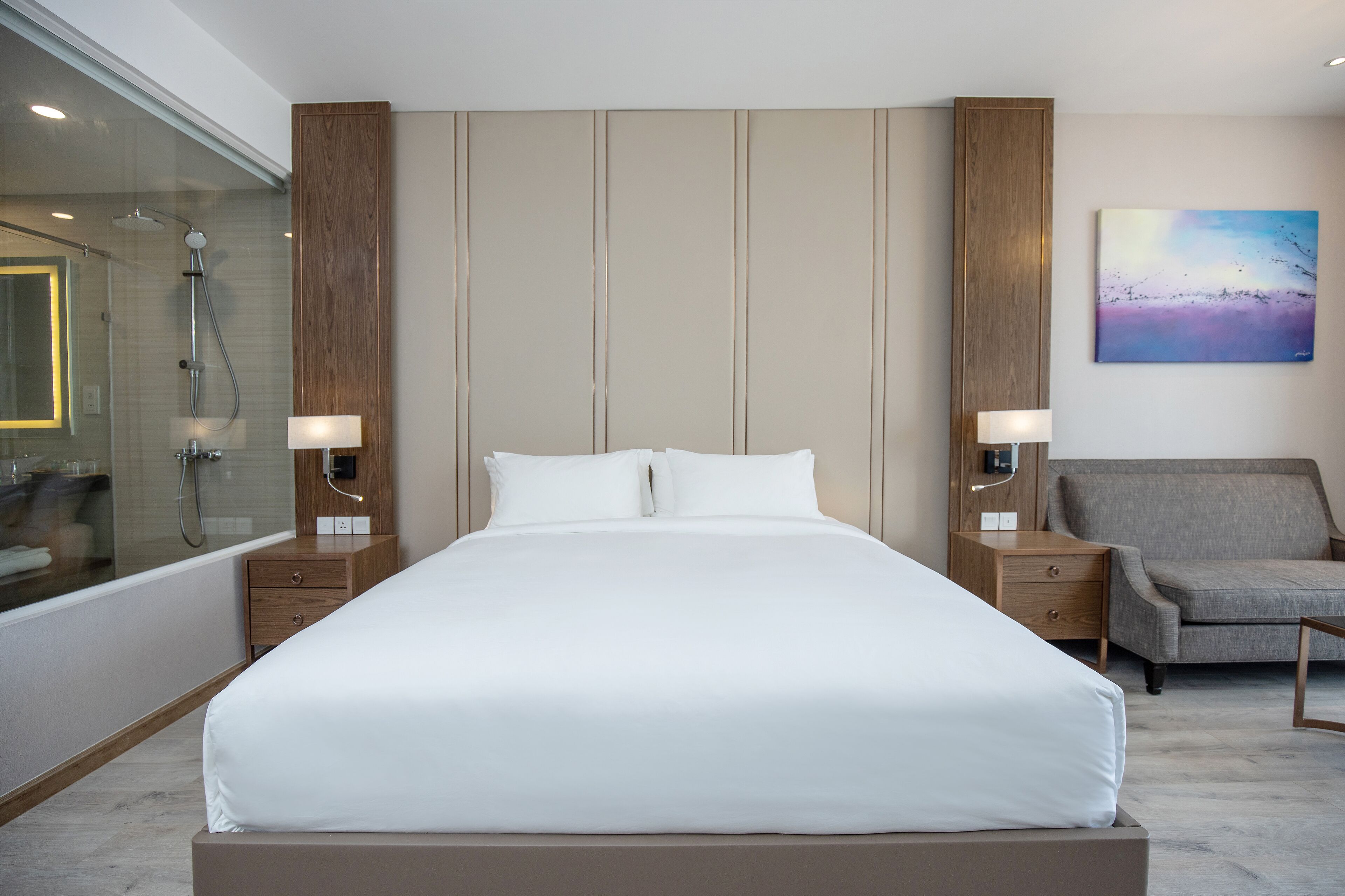 Une chambre d’hôtel moderne avec un grand lit, des tables de chevet avec des lampes, un fauteuil gris, une cabine de douche en verre et un tableau au mur.