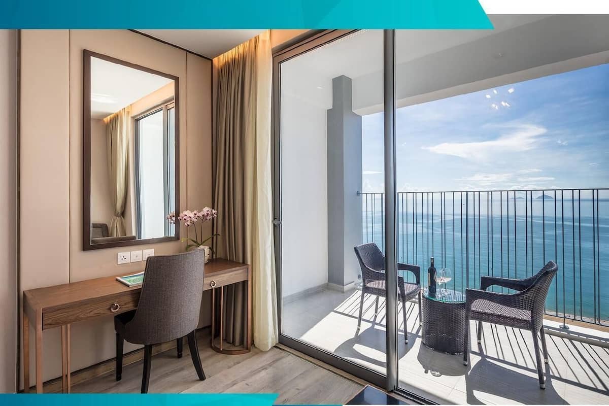 Habitación de hotel con escritorio, silla y un amplio ventanal corredero que da a un balcón con vista al mar.