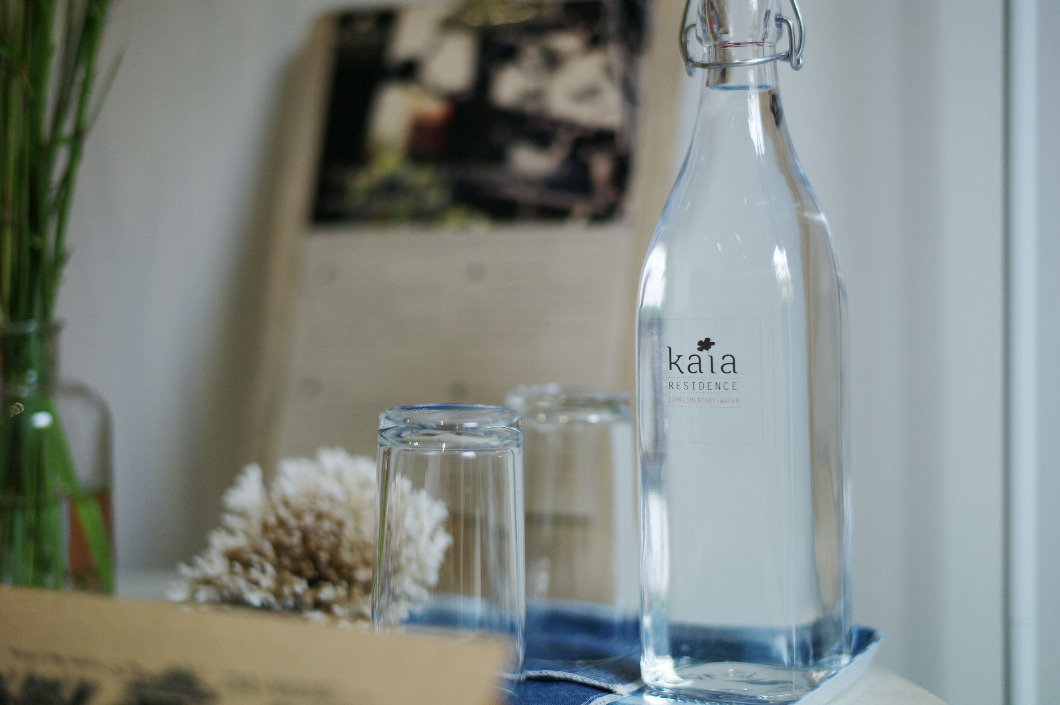 Une bouteille en verre transparent portant l’étiquette « Kaia Residence », avec un tissu bleu placé en dessous.