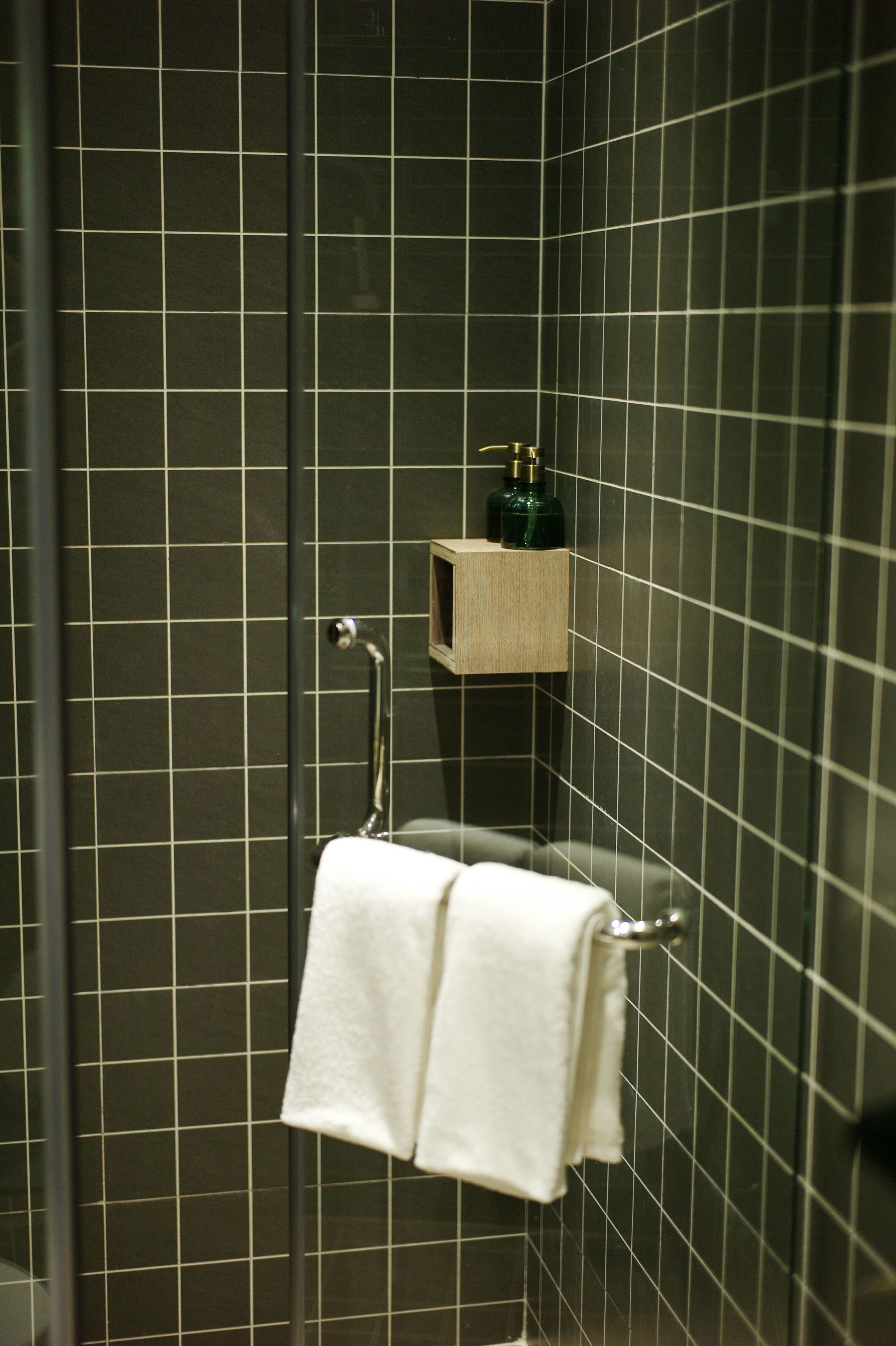 A bathroom with dark tiled walls, a glass shower enclosure, a towel rack with two white towels, and a small wooden shelf with a green bottle.