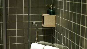 A bathroom with dark tiled walls, a glass shower enclosure, a towel rack with two white towels, and a small wooden shelf with a green bottle.