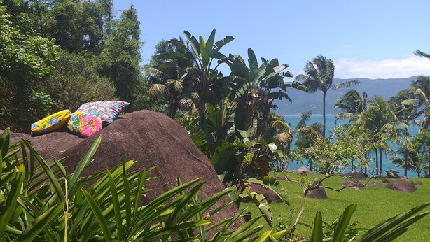 A tropical garden with a large rock, colorful pillows, and a view of the ocean and mountains. - GuestHouse Elephant Ilhabela (Urubu)