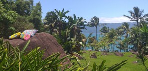 Ein tropischer Garten mit einem großen Felsen, bunten Kissen und Blick auf das Meer und die Berge.