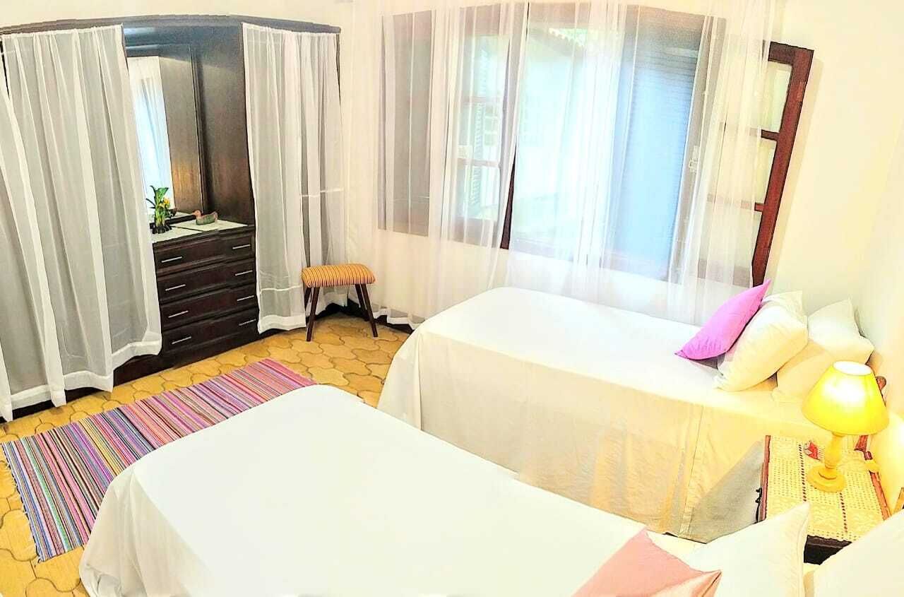 A bedroom with two beds, a dresser, a bench, a window with sheer curtains, and a bedside lamp.