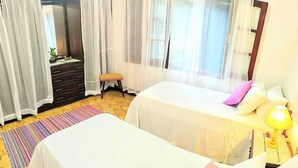 A bedroom with two beds, a dresser, a bench, a window with sheer curtains, and a bedside lamp. - GuestHouse Elephant Ilhabela (Urubu)