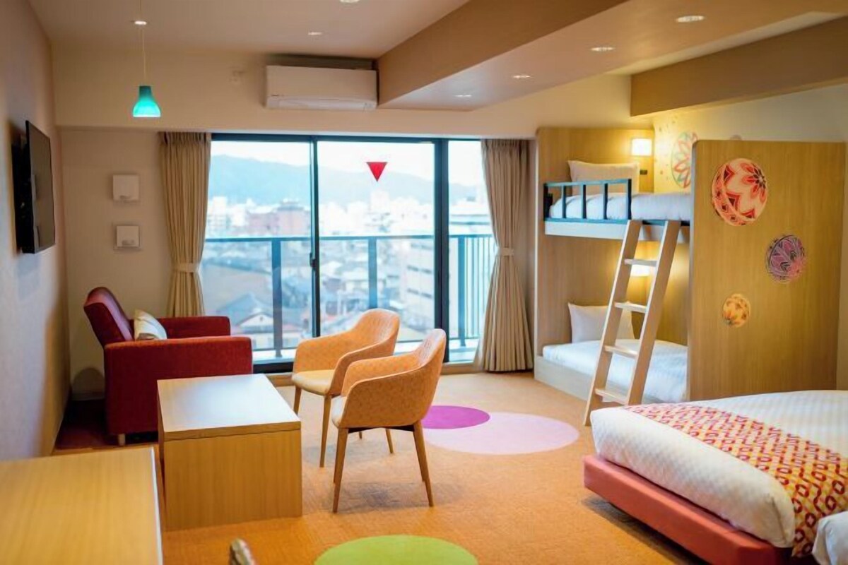 A hotel room with a bunk bed, a desk, two chairs, a red sofa, and a large window with a view of a cityscape.