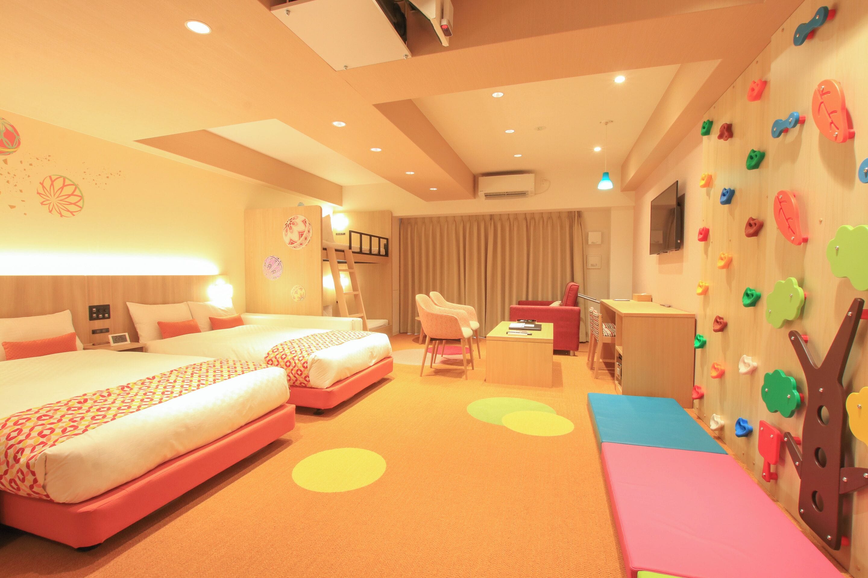 A hotel room with two beds, a desk, a chair, a TV, and a wall with colorful climbing holds.