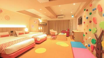 A hotel room with two beds, a desk, a chair, a TV, and a wall with colorful climbing holds.