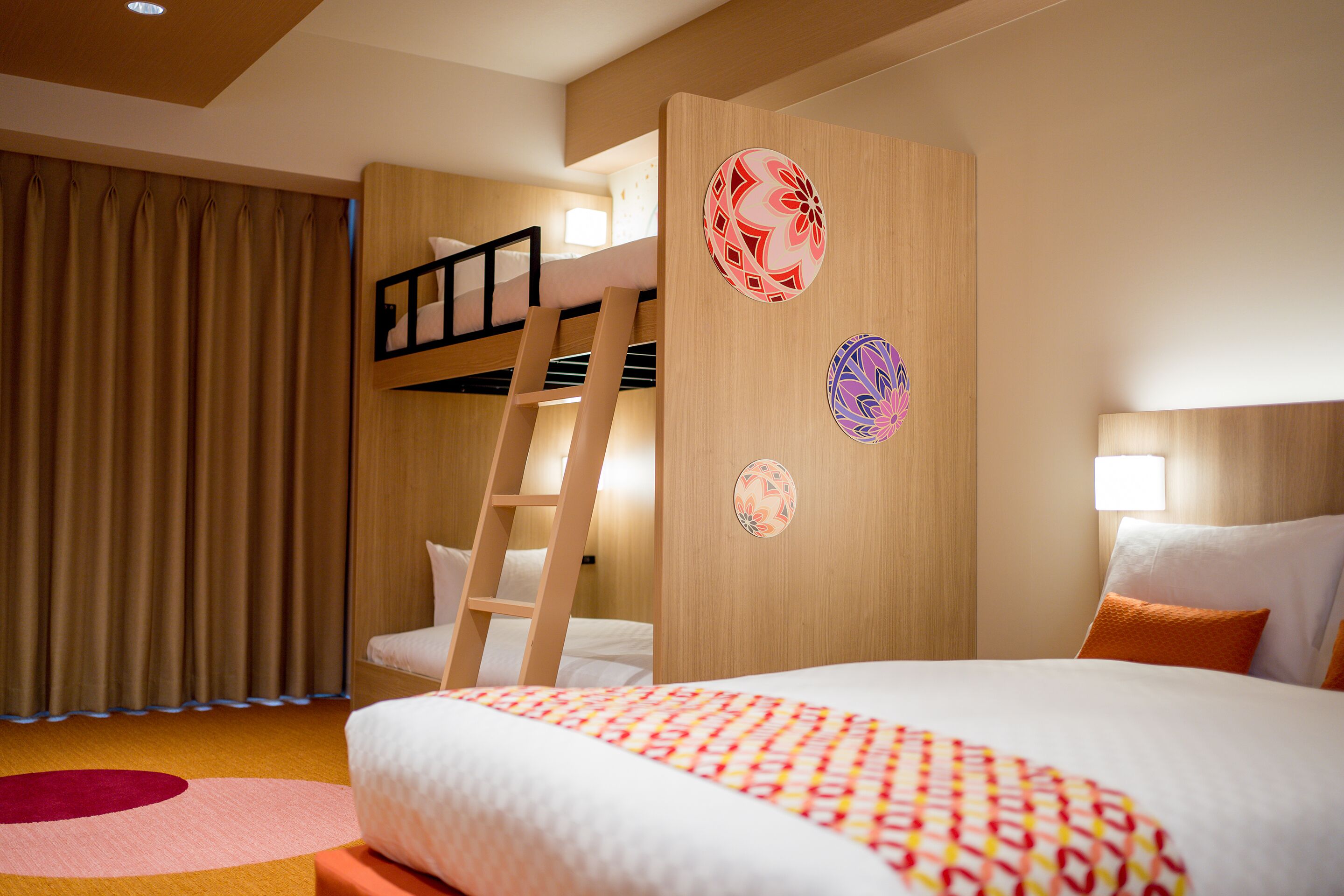 Una camera d'albergo con un letto a castello, una scala, un letto grande con copriletto fantasia e adesivi decorativi al muro.