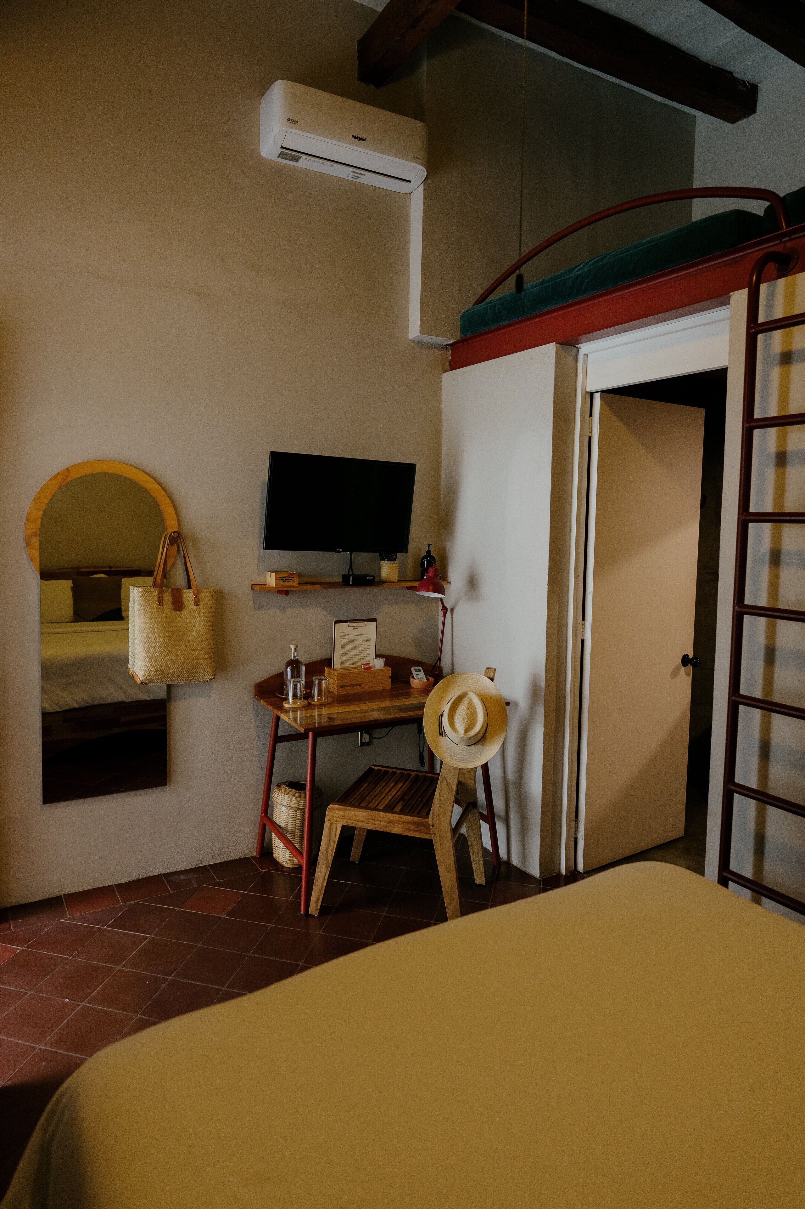 A bedroom with a bed, a desk with a chair, a television, an air conditioning unit, a wardrobe, and a door.