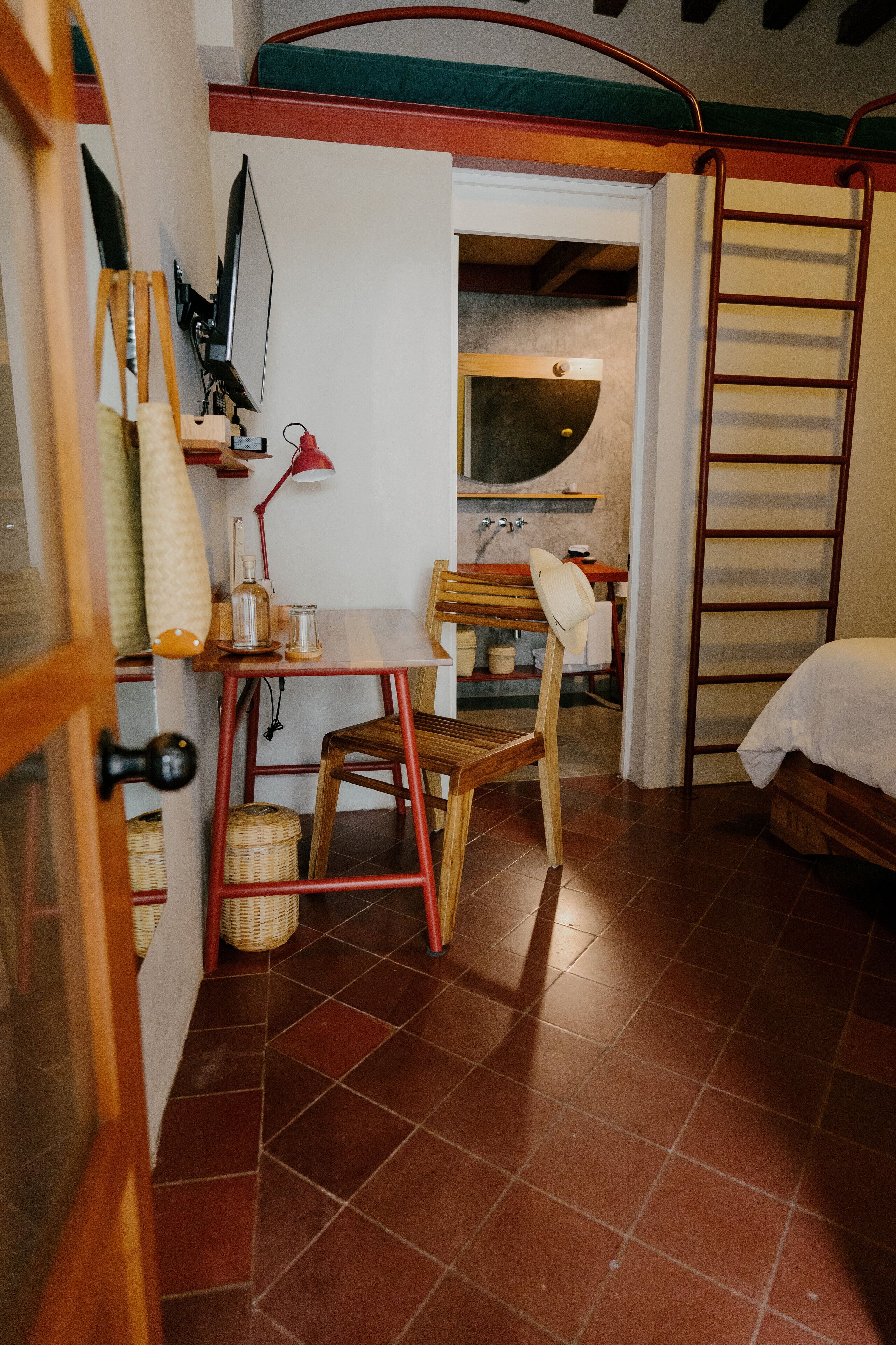 A compact room with a bed, a table with chairs, a desk with a lamp, and a ladder against the wall.