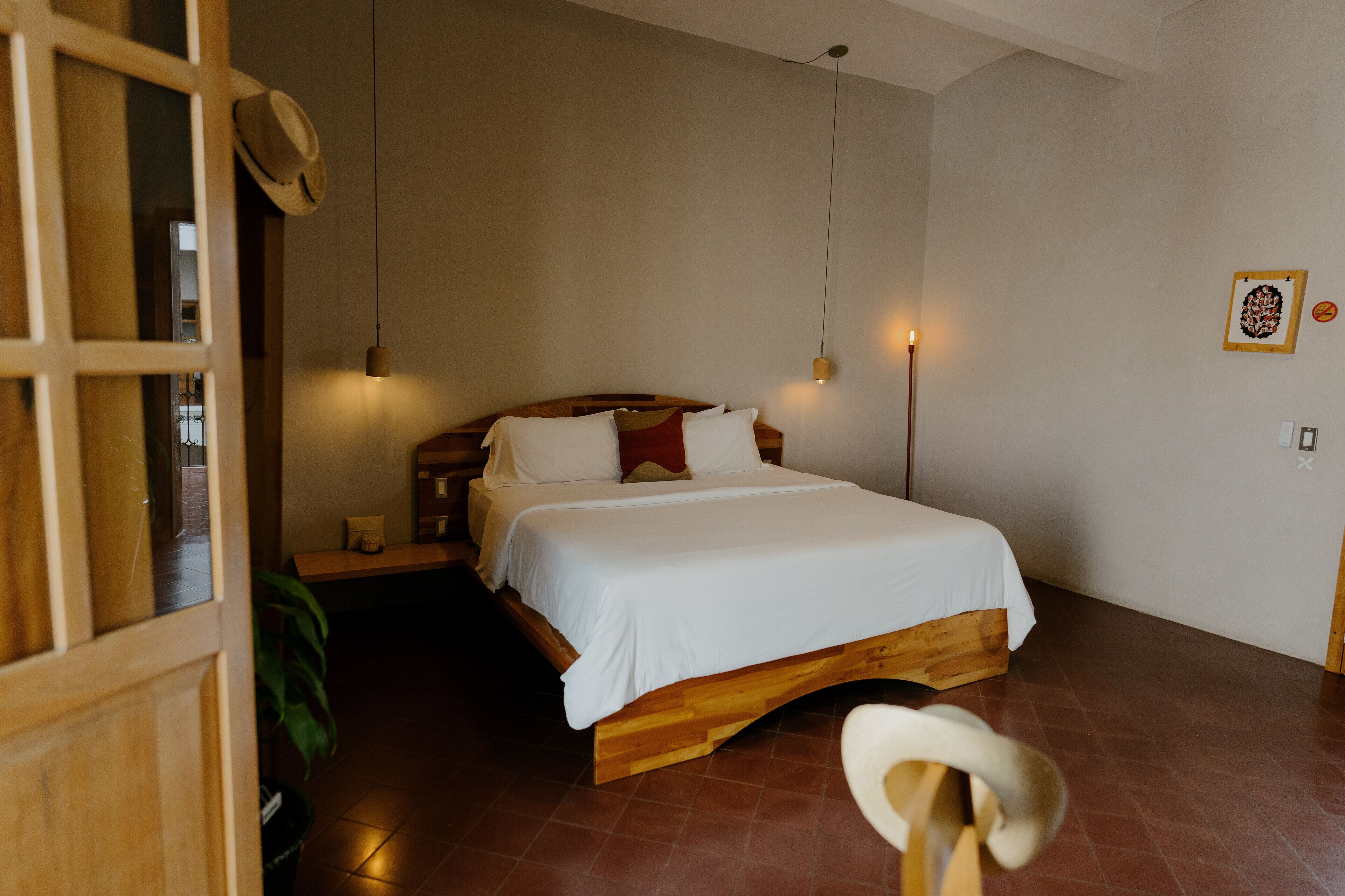 A wooden bed with white bedding, a bench, and a hanging light fixture in a room with a tiled floor.