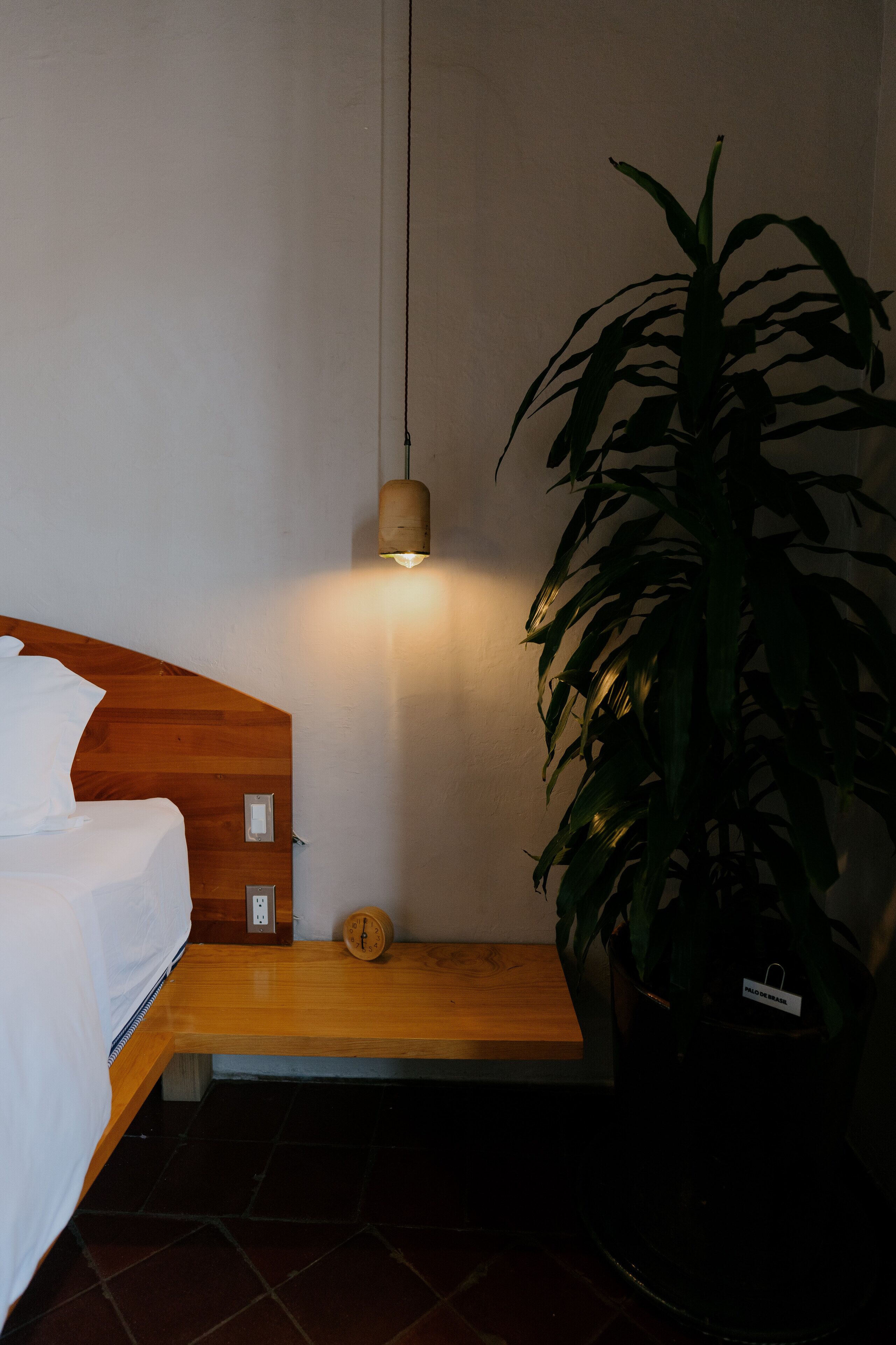 A bedroom with a wooden headboard, a bedside table, a clock, a potted plant, and a hanging light fixture.