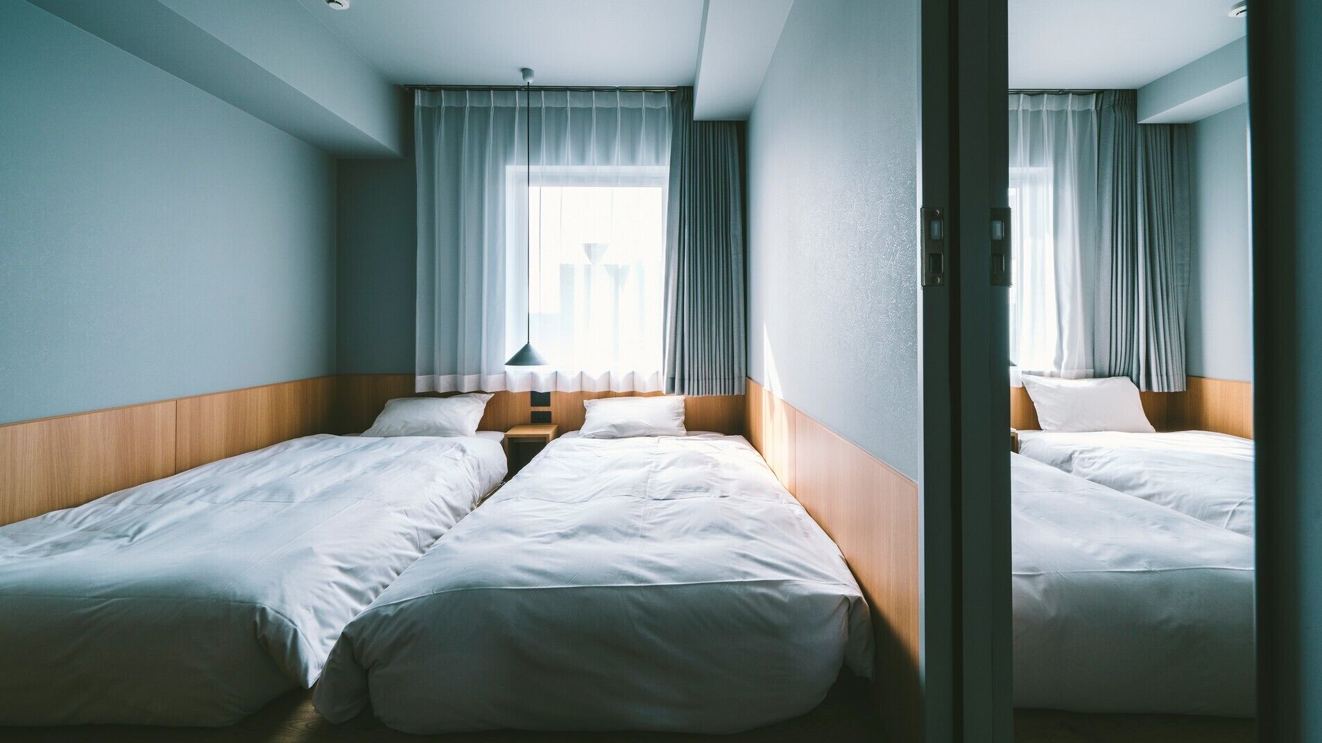 A hotel room with two single beds, a wooden headboard, a nightstand, and a window with curtains.