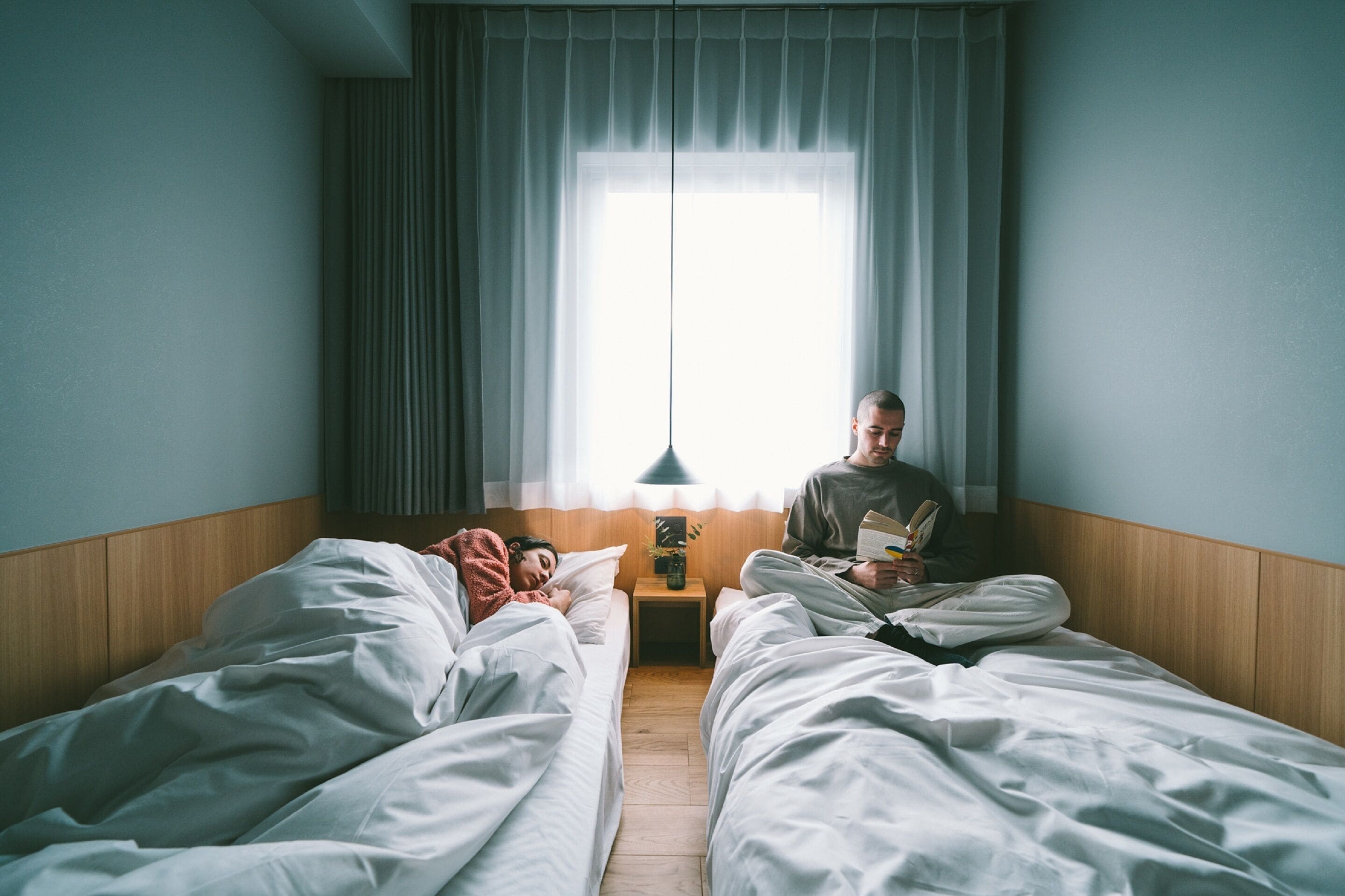 A hotel room with two beds, a window with curtains, a bedside table with a lamp, and a small plant.