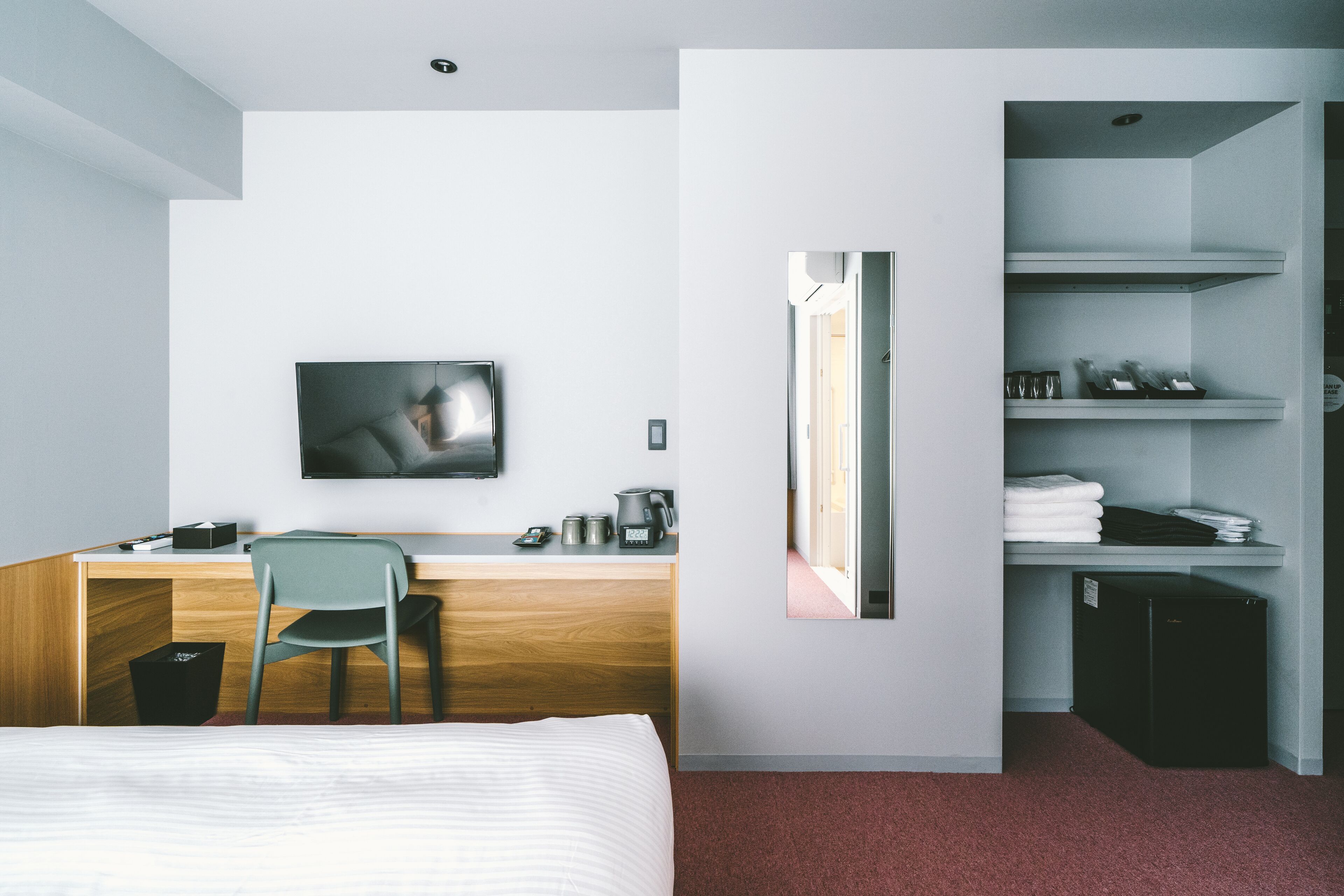 A hotel room with a bed, a desk with a chair, a television, a mirror, and a small kitchenette with a refrigerator.
