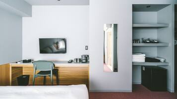A hotel room with a bed, a desk with a chair, a television, a mirror, and a small kitchenette with a refrigerator.