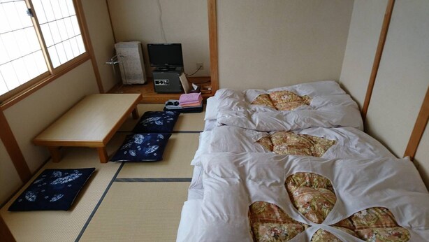 A room with tatami flooring, a futon bed, a small wooden table, a television, and a refrigerator. - Apple Inn Takasaka (Nozawaonsen)